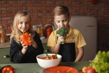 Cute girl and boy with fresh pepper and broccoli in hands standing near desk with assorted vegetables and salad bowl during vegetarian meal preparation